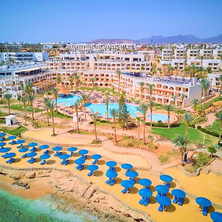 Pickalbatros Royal Grand Sharm - Adults Friendly 16 Years Plus Luaran gambar The photo shows a vibrant resort area with several buildings that appear to be accommodations or hotel units. There are multiple swimming pools surrounded by palm trees, creating a tropical atmosphere. In the foreground, there is a beach area with sa