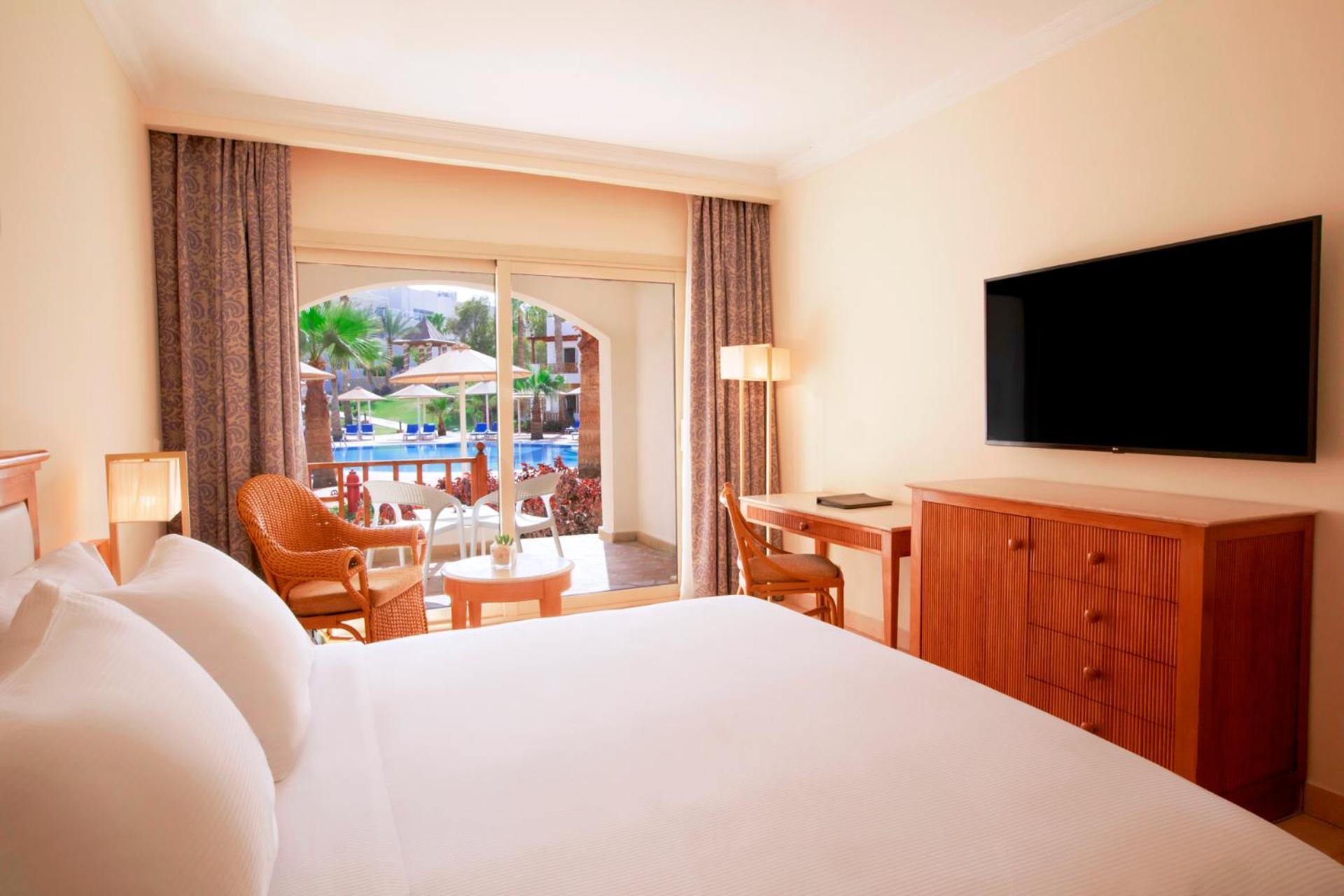 Pickalbatros Royal Grand Sharm - Adults Friendly 16 Years Plus Luaran gambar The photo shows a hotel room featuring a large bed with white bedding. There are wooden furniture pieces, including a dresser and a desk. A flat-screen television is mounted on the wall opposite the bed. The room has large windows with sheer curtains