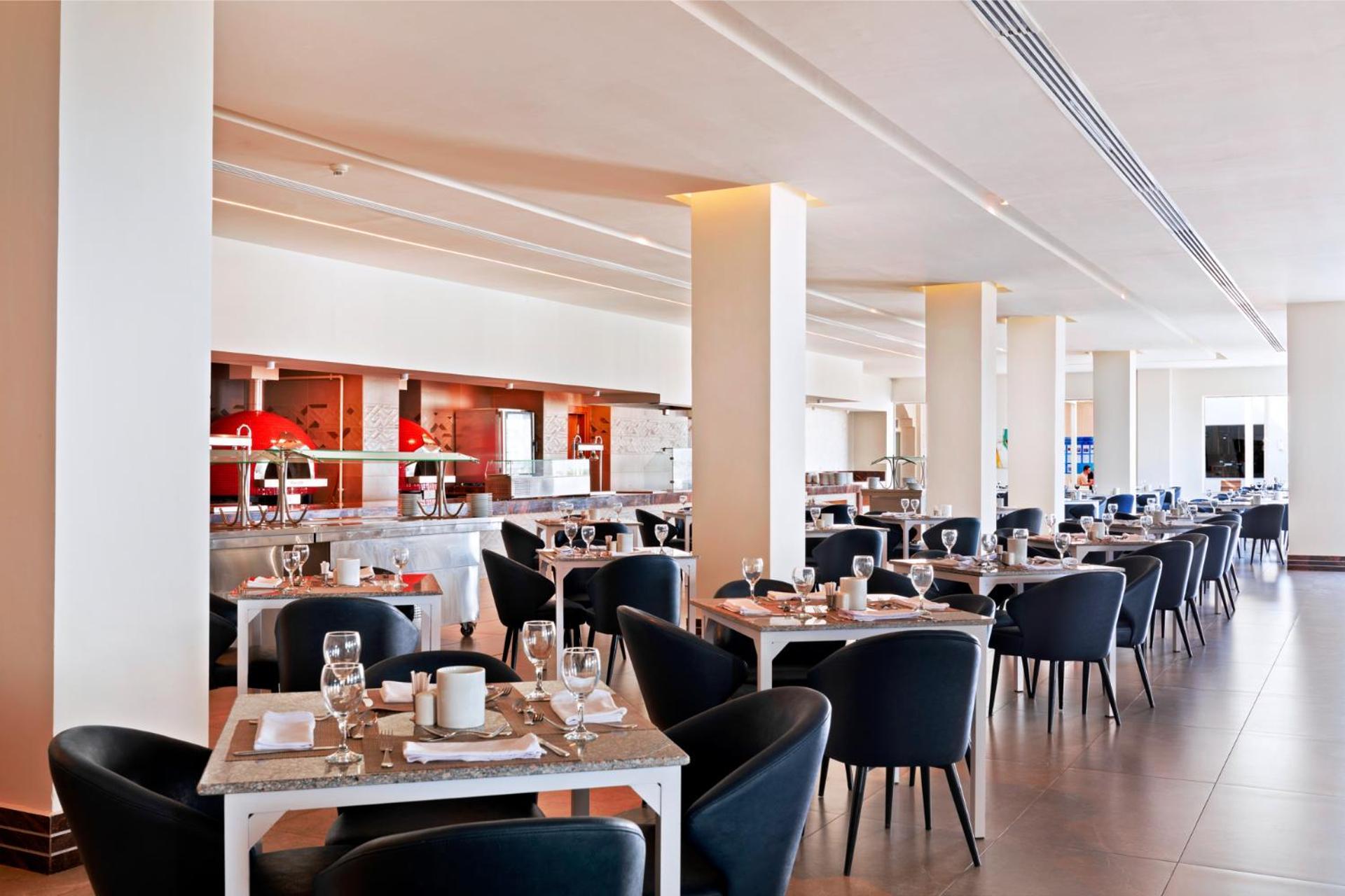 Pickalbatros Royal Grand Sharm - Adults Friendly 16 Years Plus Luaran gambar The photo shows a modern restaurant interior. It features a spacious layout with several dining tables arranged in a row, each set with dishes, glasses, and cutlery. The tables have light-colored surfaces and are accompanied by black chairs. In the b