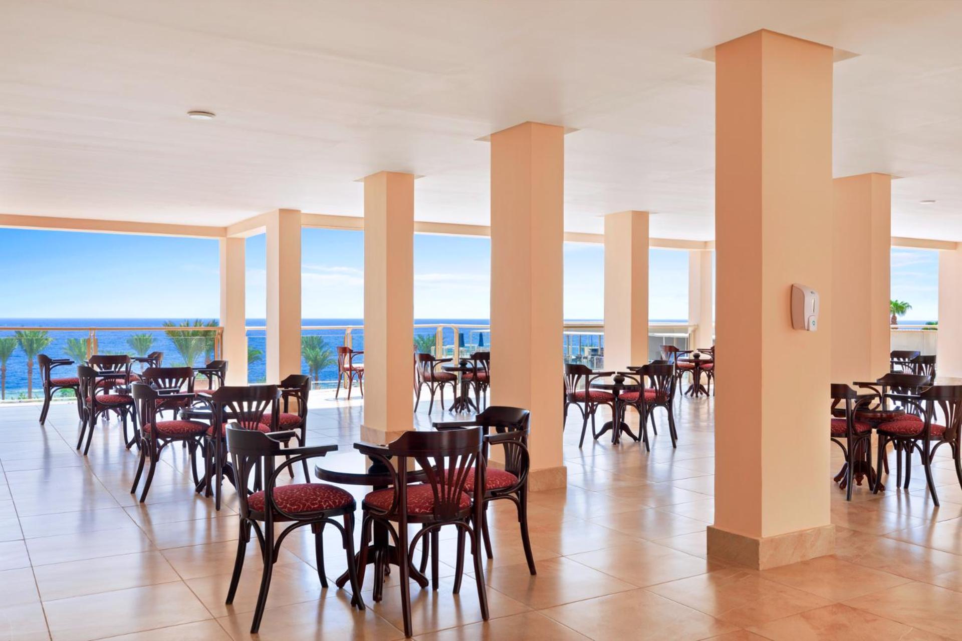 Pickalbatros Royal Grand Sharm - Adults Friendly 16 Years Plus Luaran gambar The image shows a dining area or café with several round wooden tables and chairs, featuring red upholstery. The space is well-lit, with large windows that allow a view of the ocean in the background, contributing to a bright and open atmosphere. The