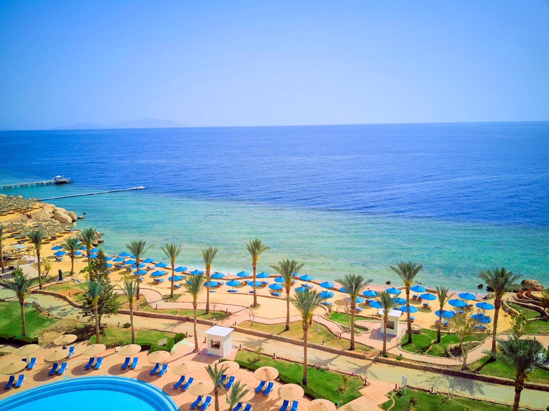 Pickalbatros Royal Grand Sharm - Adults Friendly 16 Years Plus Luaran gambar The photo displays a beautiful coastal scene featuring a clear blue sea that transitions into green tones near the shore. In the foreground, there are sandy beaches lined with palm trees and a variety of beach umbrellas. There is also a swimming pool
