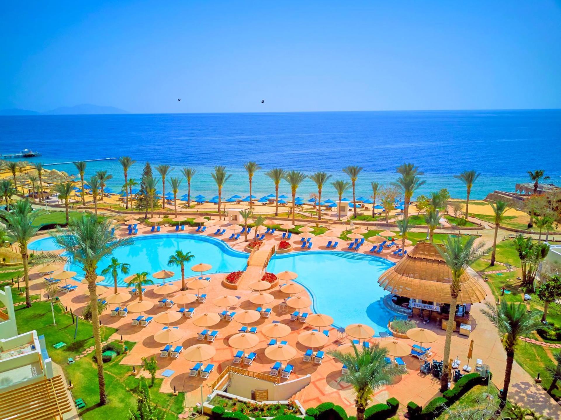 Pickalbatros Royal Grand Sharm - Adults Friendly 16 Years Plus Luaran gambar The photo depicts a picturesque resort scene by the sea. In the foreground, there is a large, inviting swimming pool surrounded by lounge chairs and umbrellas. Palm trees are strategically placed around the pool area, adding to the tropical atmospher
