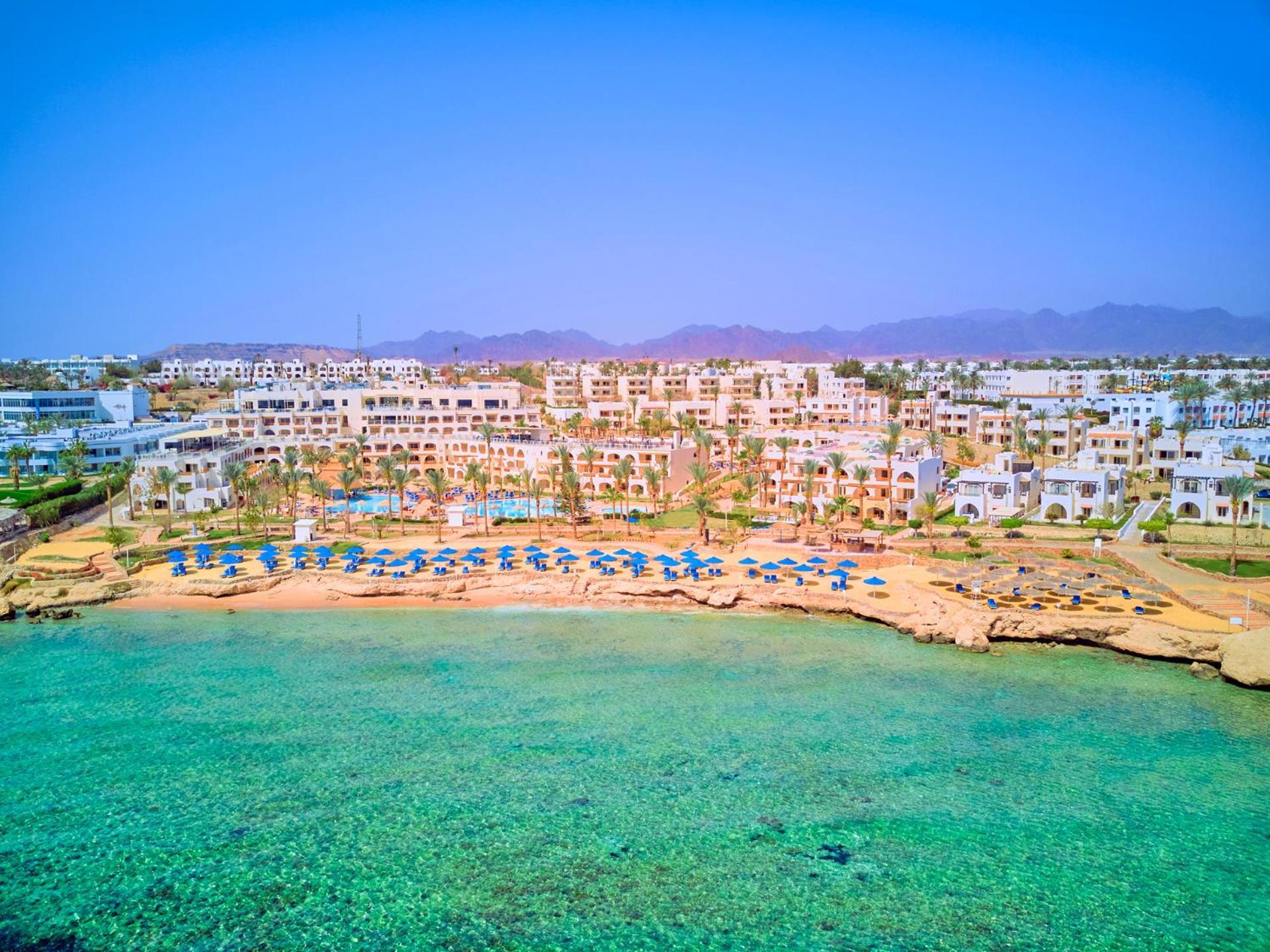 Pickalbatros Royal Grand Sharm - Adults Friendly 16 Years Plus Luaran gambar The photo depicts a picturesque beachfront resort. It shows a clear turquoise sea with a sandy shore lined with beach umbrellas and sun loungers. Behind the beach, there are several white buildings of the resort, surrounded by lush greenery. In the b