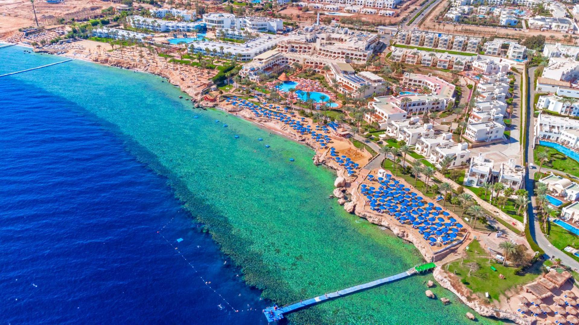 Pickalbatros Royal Grand Sharm - Adults Friendly 16 Years Plus Luaran gambar The photo showcases a beautiful aerial view of a coastal resort. In the foreground, there's a sandy beach with beach umbrellas and lounge chairs arranged neatly, indicating a tourist area. The clear, turquoise water transitions from the sandy shore t