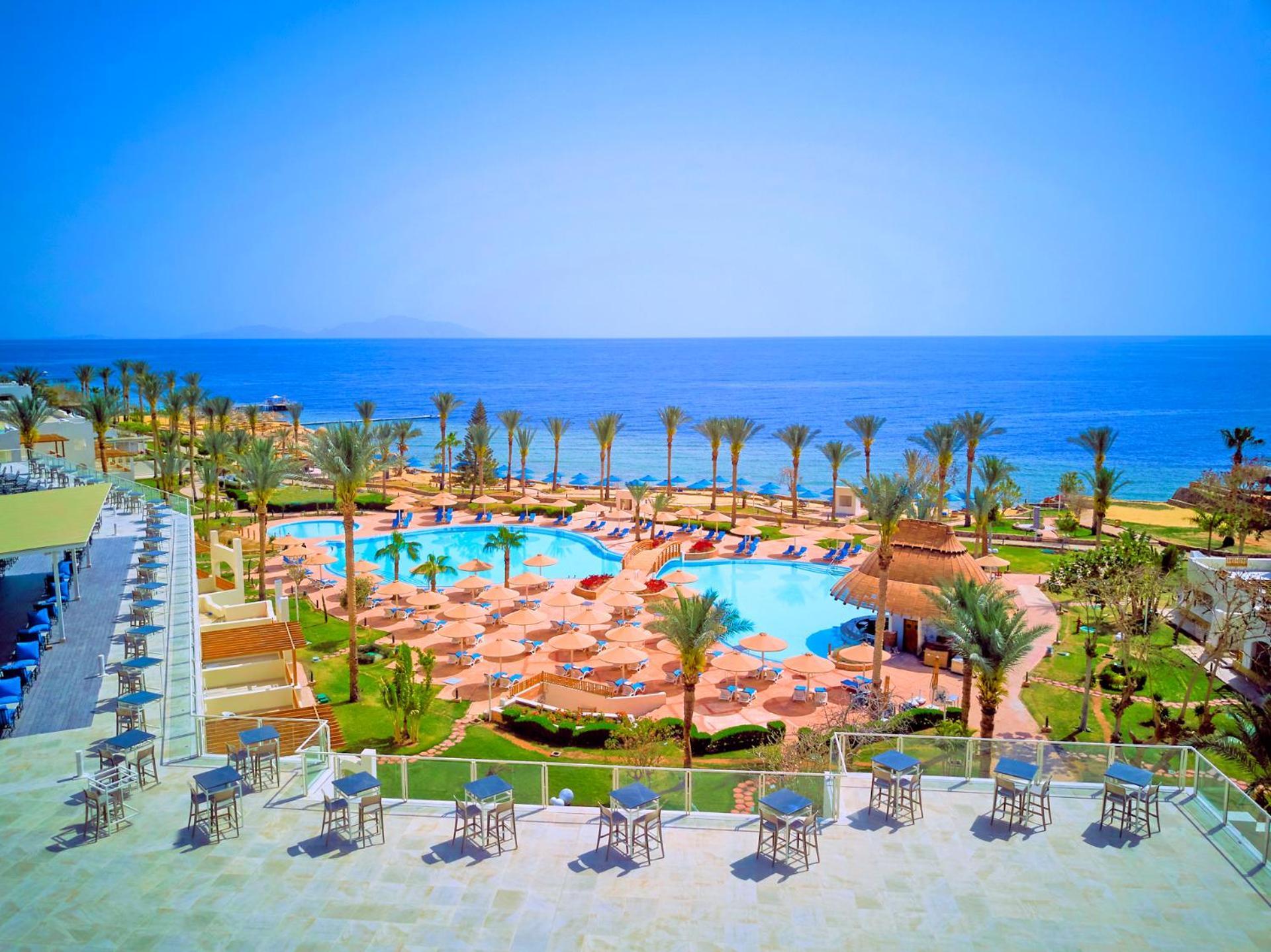 Pickalbatros Royal Grand Sharm - Adults Friendly 16 Years Plus Luaran gambar The photo shows a beautiful resort scene with a spacious swimming pool at the center, surrounded by sun loungers and palm trees. There are areas for relaxation with tables and chairs positioned around the pool. In the background, the vibrant blue oce
