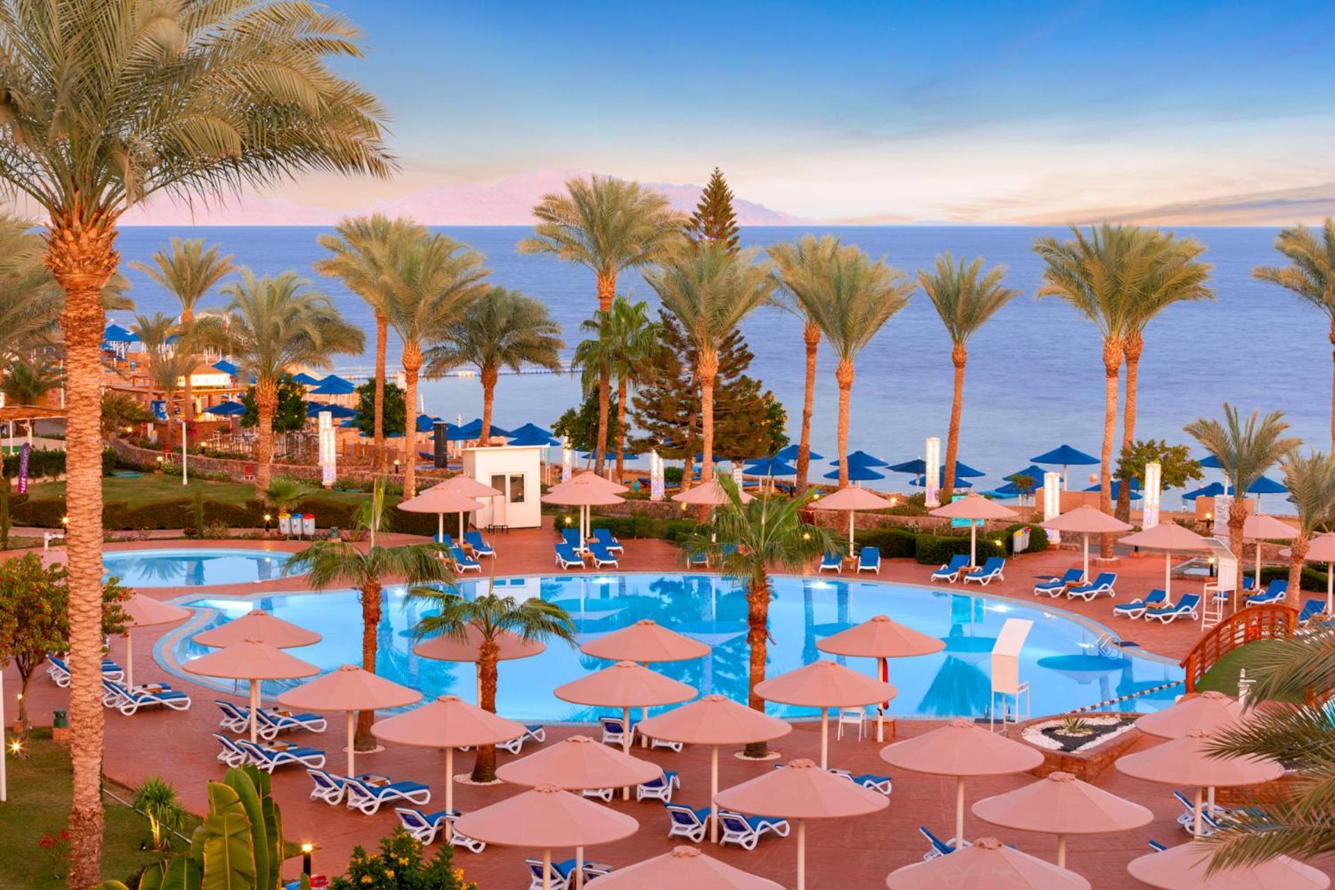 Pickalbatros Royal Grand Sharm - Adults Friendly 16 Years Plus Luaran gambar The photo shows a beautiful resort scene with a swimming pool surrounded by lounge chairs and umbrellas. Palm trees line the area, adding to the tropical atmosphere. In the background, there is a view of the sea under a clear sky, suggesting a peacef
