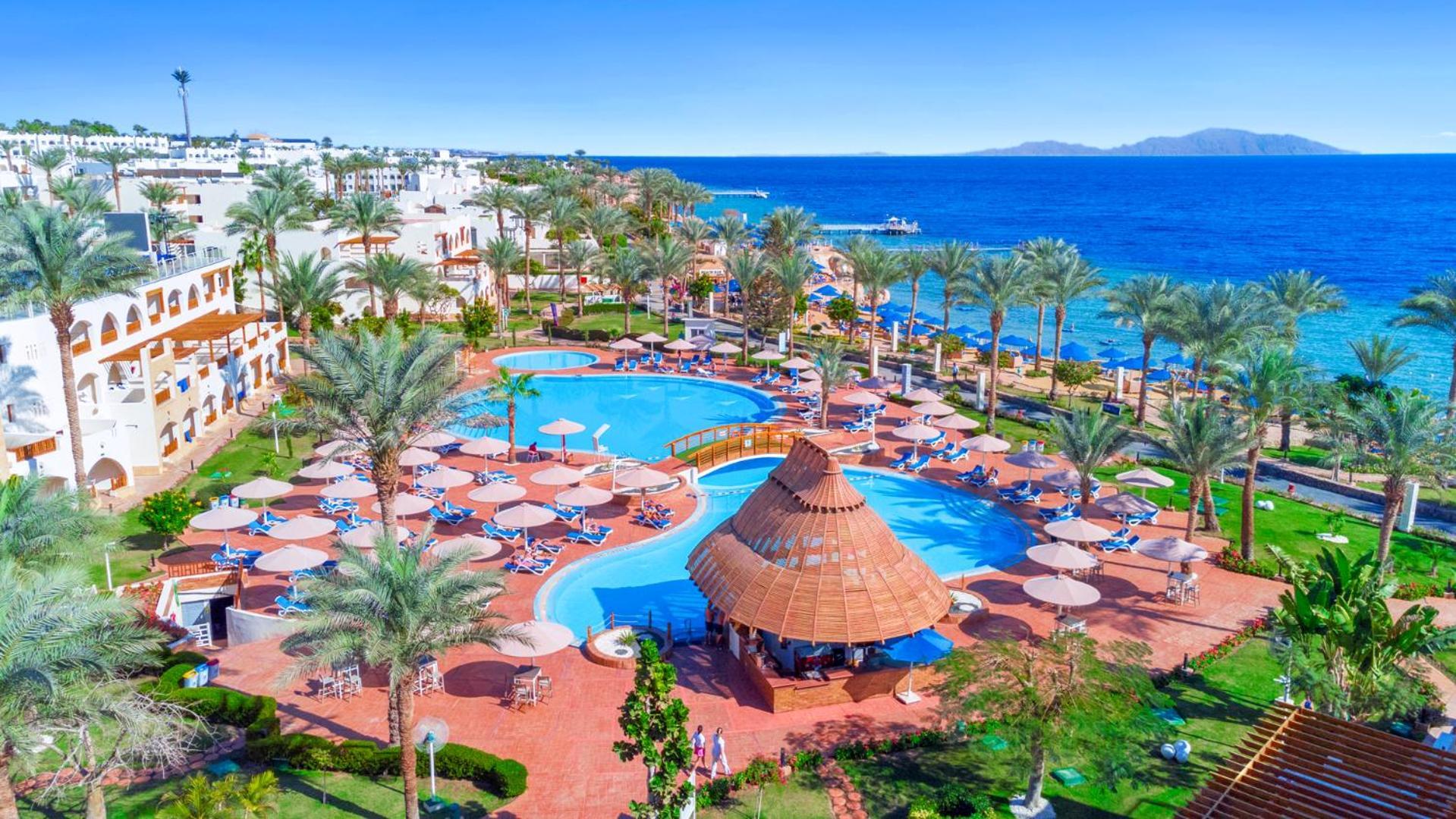 Pickalbatros Royal Grand Sharm - Adults Friendly 16 Years Plus Luaran gambar The image depicts a vibrant resort scene overlooking a clear blue sea. In the foreground, there are multiple swimming pools surrounded by sun loungers and umbrellas. A thatched-roof gazebo is situated near the center of the pool area. Palms and lush 