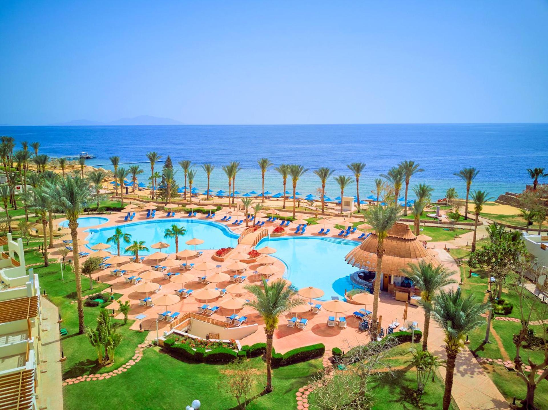 Pickalbatros Royal Grand Sharm - Adults Friendly 16 Years Plus Luaran gambar The photo depicts a beautiful resort scene by the ocean. In the foreground, there is a large swimming pool surrounded by palm trees and lounge chairs. The pool area features sunbeds and umbrellas, suggesting a relaxing atmosphere for guests. In the b