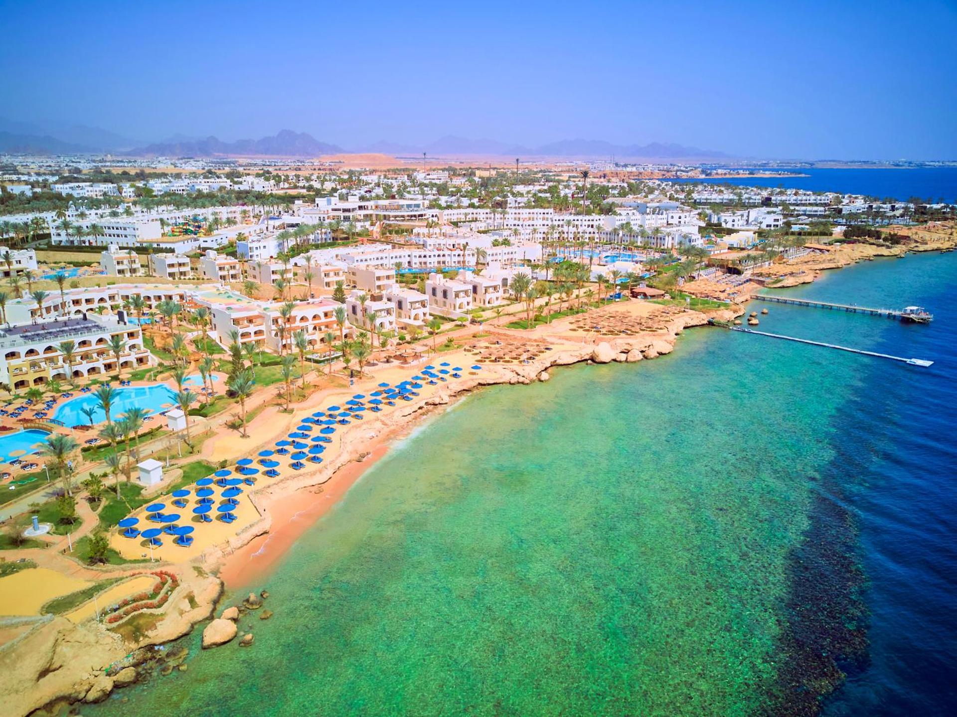 Pickalbatros Royal Grand Sharm - Adults Friendly 16 Years Plus Luaran gambar The photo showcases a picturesque coastal scene, likely from a resort area. It features a sandy beach lined with blue umbrellas and lounge chairs, indicating a recreational space for relaxation. The water is clear and exhibits shades of green and blu