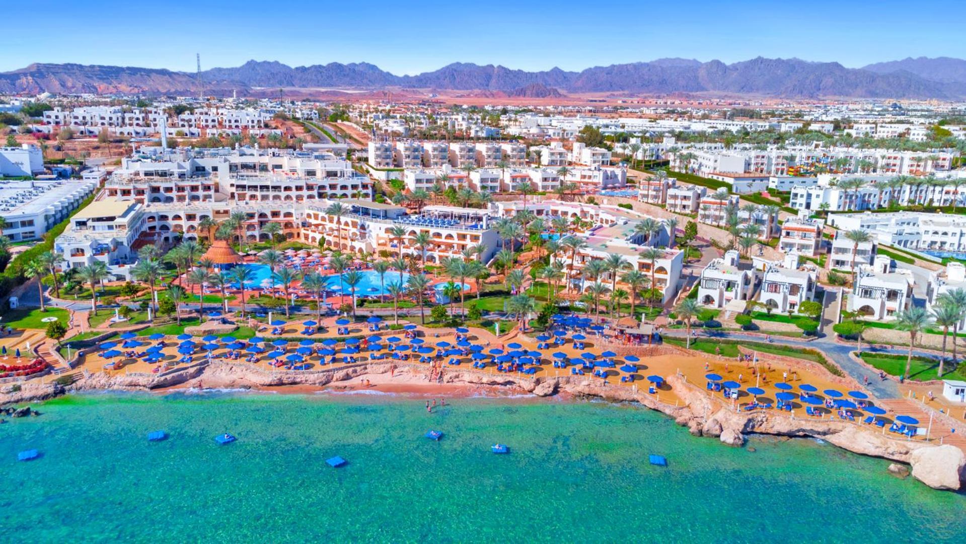 Pickalbatros Royal Grand Sharm - Adults Friendly 16 Years Plus Luaran gambar The photo showcases a beautiful coastal resort area. In the foreground, there is a sandy beach lined with blue umbrellas and sun loungers. The vibrant turquoise water gently laps at the shore. Behind the beach, you can see a well-maintained resort wi
