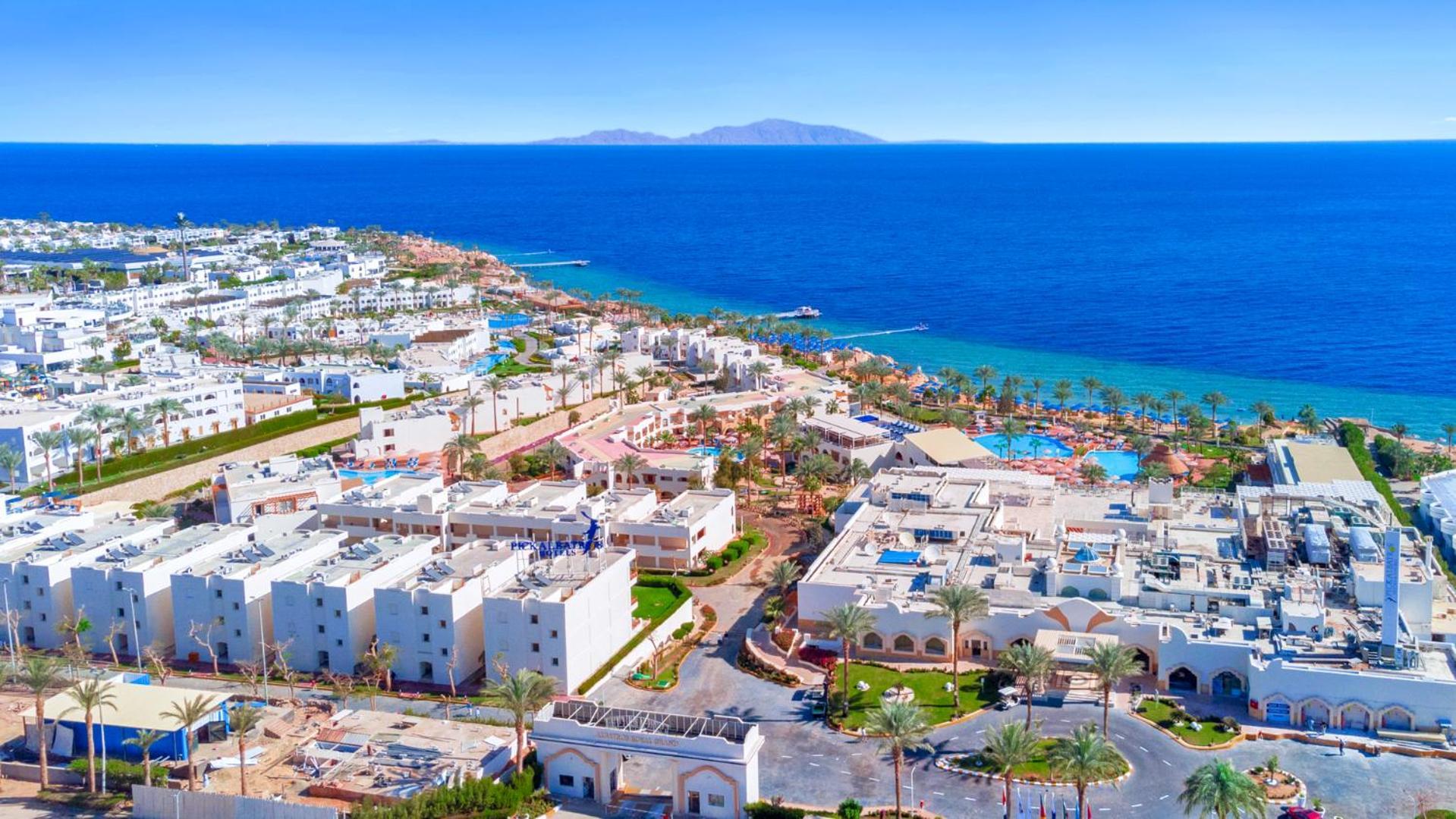 Pickalbatros Royal Grand Sharm - Adults Friendly 16 Years Plus Luaran gambar The photo shows a beautiful coastal landscape, featuring a resort area by the sea. There are white buildings, likely hotels or villas, arranged in a sprawling layout near the beach. The water is a vibrant blue, contrasting with the sandy shore and gr