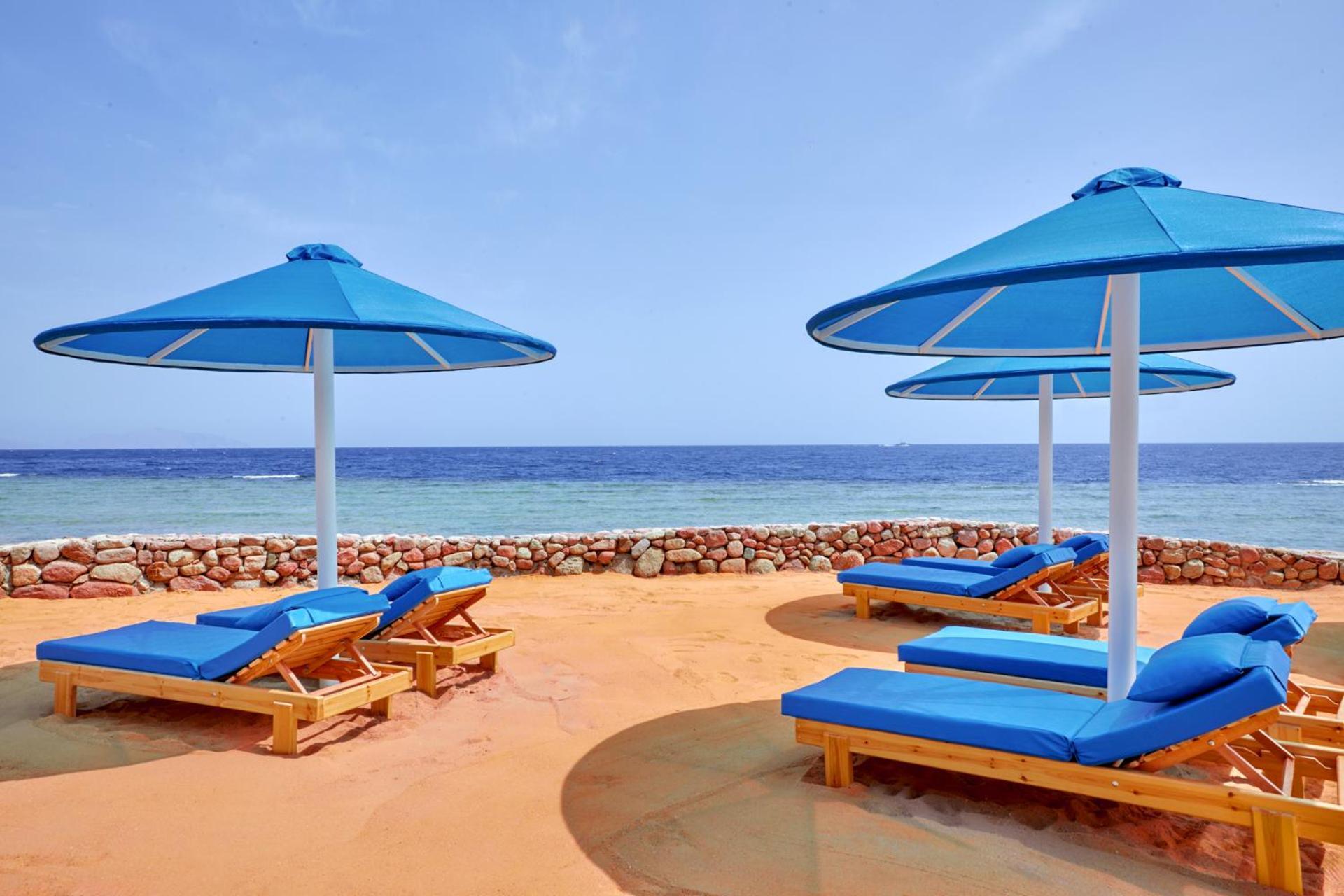 Pickalbatros Royal Grand Sharm - Adults Friendly 16 Years Plus Luaran gambar The photo shows a beach scene featuring several blue umbrellas and lounge chairs on a sandy area. The umbrellas are large and have a bright blue color, providing shade for the lounge chairs underneath. In the background, there's a view of the ocean w