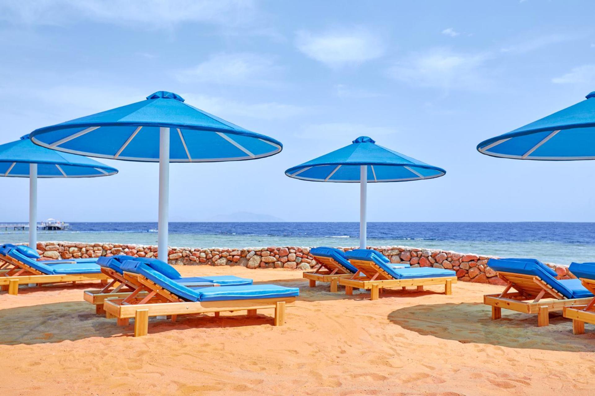 Pickalbatros Royal Grand Sharm - Adults Friendly 16 Years Plus Luaran gambar The photo shows a beach scene featuring several blue umbrellas providing shade over wooden lounge chairs. The sandy beach extends toward a calm sea, creating a relaxing atmosphere. The sky is mostly clear, with a few clouds, suggesting pleasant weath