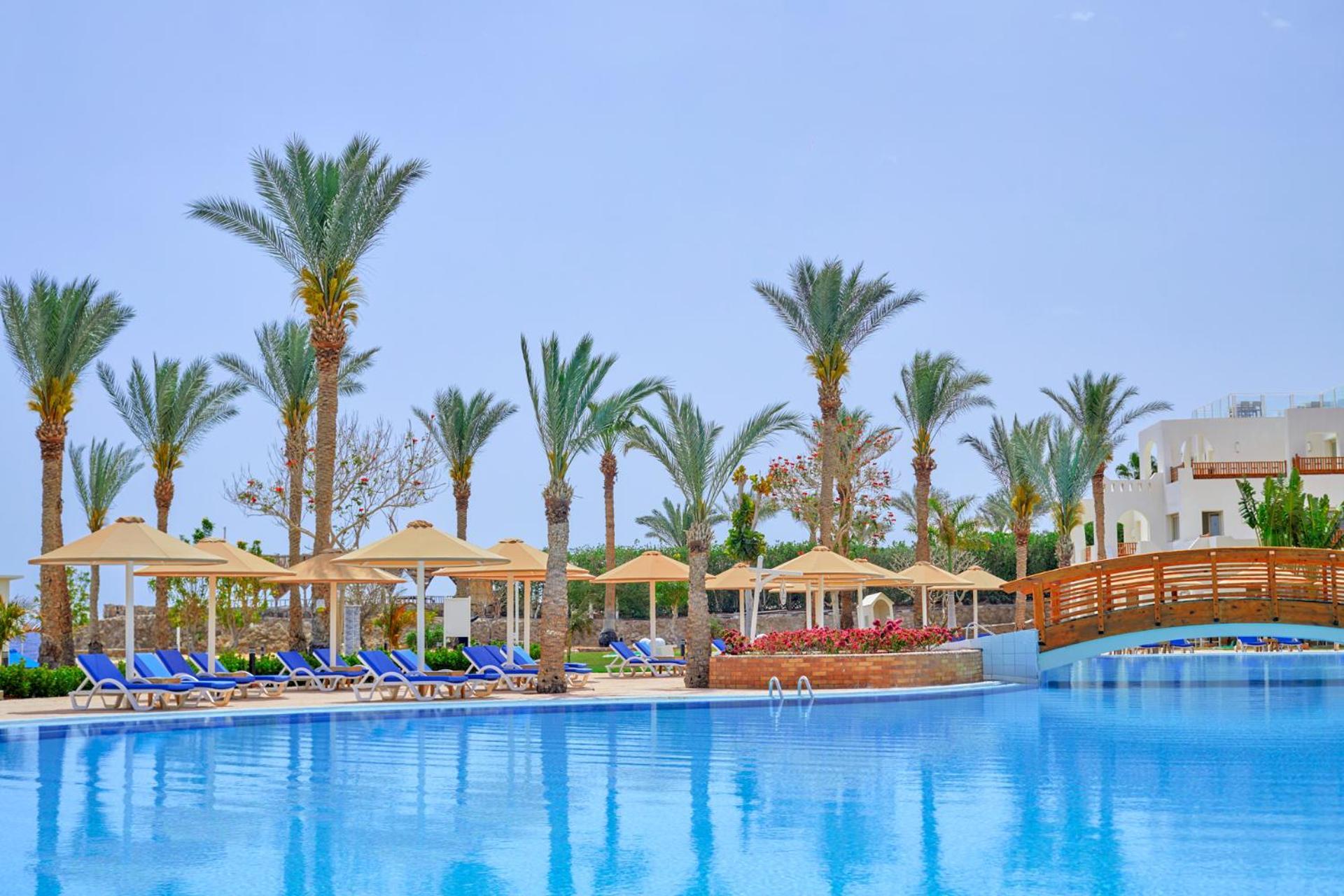 Pickalbatros Royal Grand Sharm - Adults Friendly 16 Years Plus Luaran gambar The photo shows a serene poolside scene at a resort. There is a large, clear blue pool reflecting the sky, surrounded by lounge chairs with blue cushions. Palm trees line the area, adding a tropical feel. Several covered loungers are placed under umb