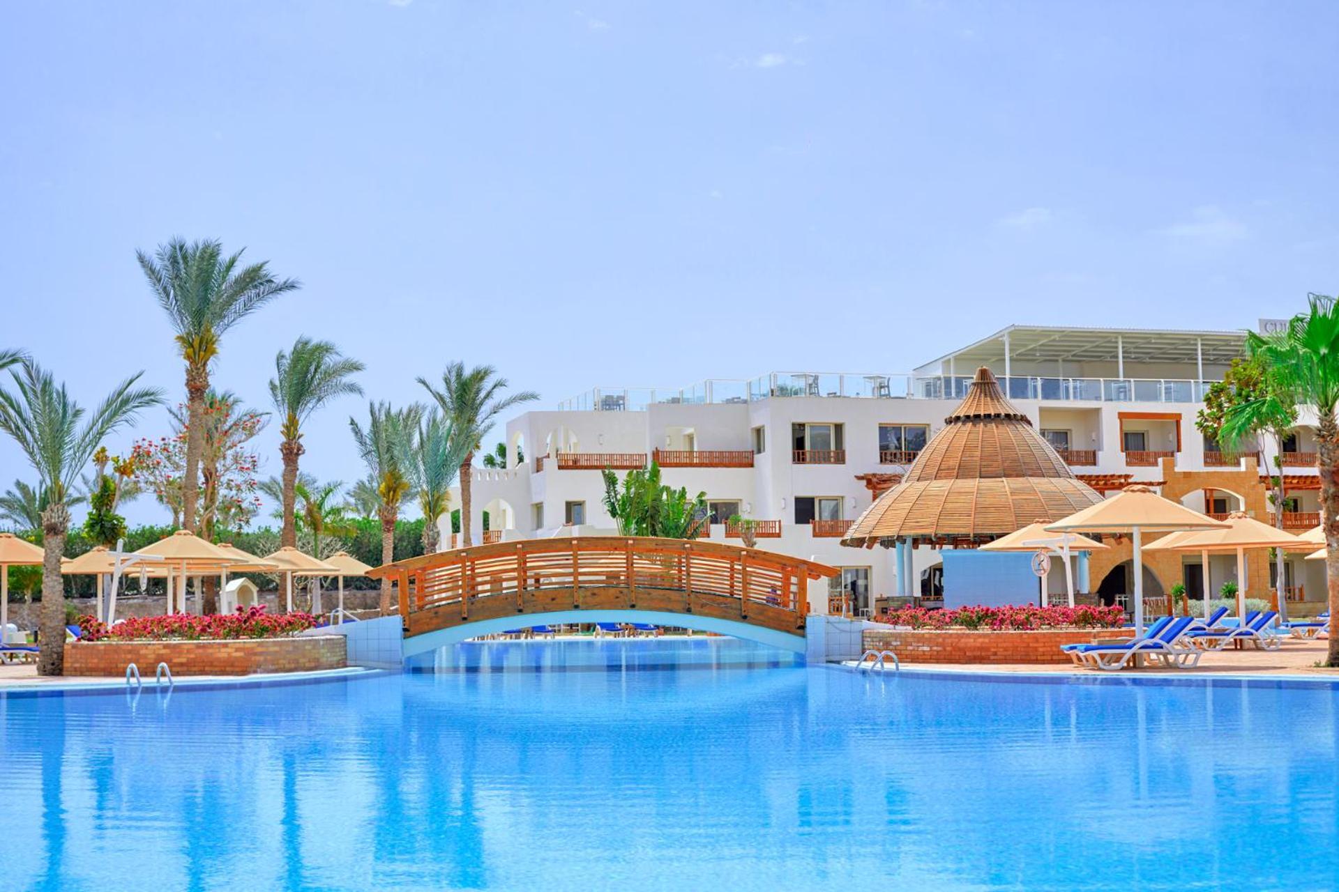 Pickalbatros Royal Grand Sharm - Adults Friendly 16 Years Plus Luaran gambar The photo depicts a serene resort setting featuring a large, calm swimming pool, which reflects the clear blue sky. In the foreground, a wooden bridge arches over the pool. Surrounding the pool are several palm trees and tropical foliage, contributin