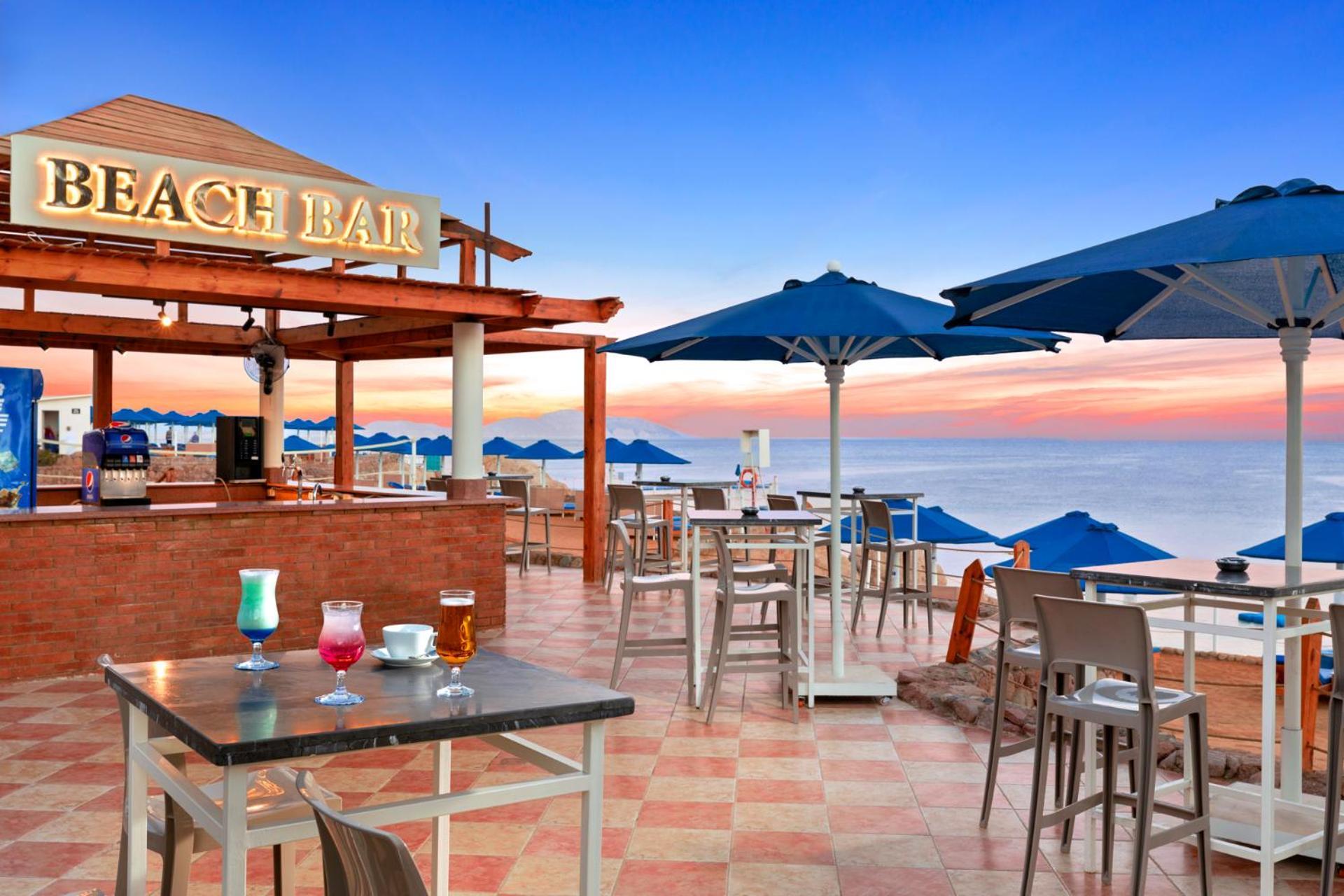 Pickalbatros Royal Grand Sharm - Adults Friendly 16 Years Plus Luaran gambar The photo shows a beach bar setting, featuring a wooden structure with the words "BEACH BAR" prominently displayed. There are several tables and chairs arranged on a tiled floor, with some drinks placed on the tables. Blue umbrellas provide shade for