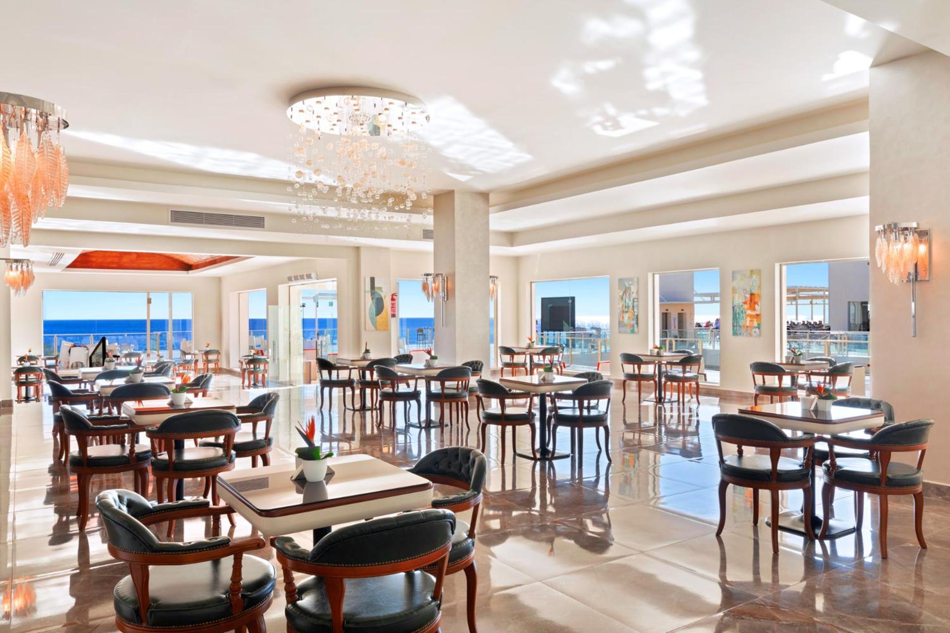 Pickalbatros Royal Grand Sharm - Adults Friendly 16 Years Plus Luaran gambar The photo shows a bright and elegant restaurant interior. There are several tables arranged throughout the space, each with chairs around them. The decor features modern lighting, including a chandelier, and large windows that allow natural light to 