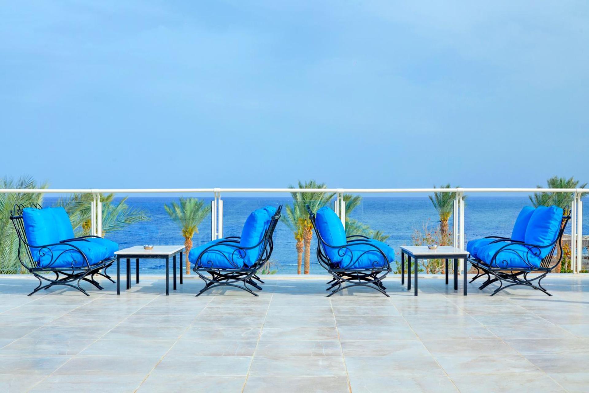 Pickalbatros Royal Grand Sharm - Adults Friendly 16 Years Plus Luaran gambar The photo depicts a serene outdoor patio area with four blue lounge chairs arranged around small tables. The setting features a clear view of the ocean with palm trees visible in the background, complementing the tranquil atmosphere. The overall colo