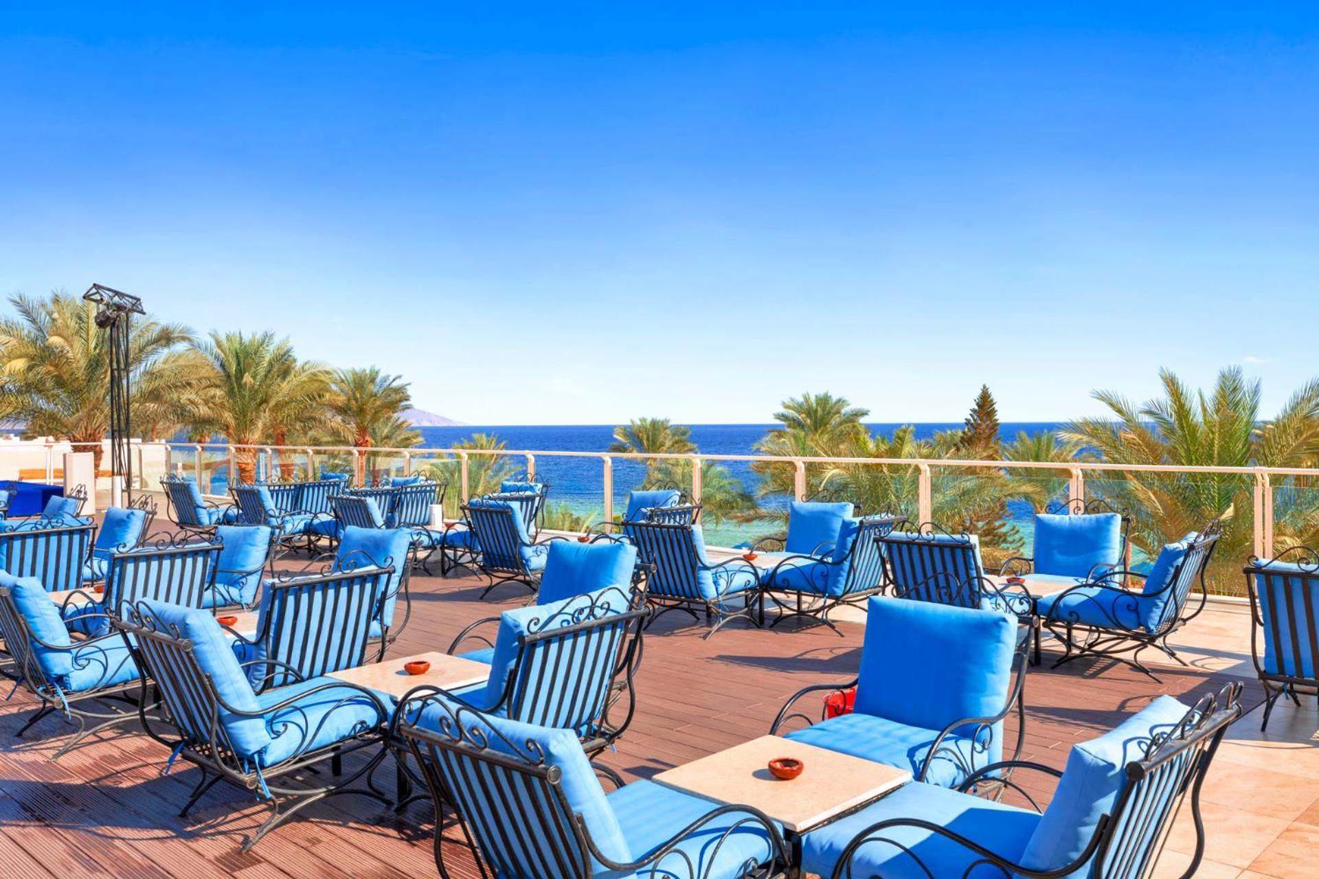 Pickalbatros Royal Grand Sharm - Adults Friendly 16 Years Plus Luaran gambar The photo shows an outdoor deck or terrace area, adorned with numerous blue lounge chairs arranged neatly. In the background, there are palm trees and a clear blue sea that stretches toward the horizon under a bright sky. The setting appears to be a 